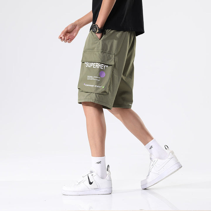 Un homme portant un short cargo ample léger avec inscriptions, kaki. Tailles disponibles du M au 8XL. Idéal pour les journées chaudes.