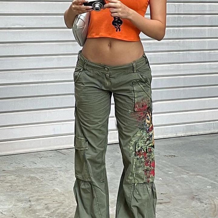 Une femme pose devant un rideau de magasin dans la rue. Elle porte un top orange court, un sac et un pantalon cargo taille basse évasée kaki avec un motif coloré sur l'une des jambes. Elle a dans ses mains un appareil photo et des clés.