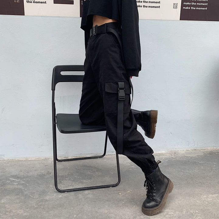 Une personne portant un pantalon noir et une chemise noire, assise sur une chaise, avec un zoom sur une botte noire. Pantalon cargo resserré aux chevilles poches clip - Noir - Femme.