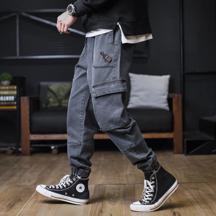 Un homme portant des sneakers noires et un pantalon gris, avec un jean cargo jogger gris foncé et une poche ornée d'un patch guitare.