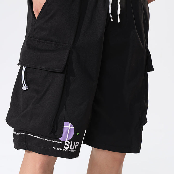 Un homme porte un short cargo ample noir avec inscriptions, idéal pour un style décontracté. Ce short en polyester léger est disponible en différentes tailles.
