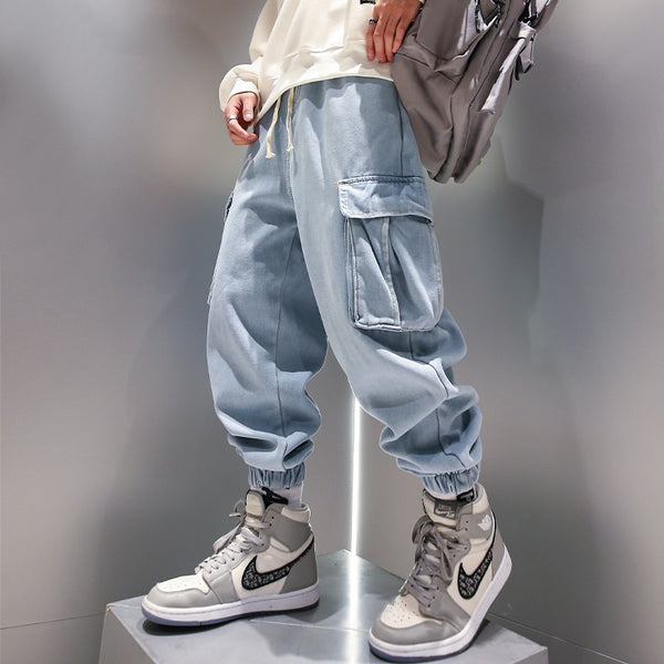 Un homme pose devant un mur gris sur un socle gris. Il porte un pull blanc, des baskets blanches et grises ainsi qu'un jean baggy resserré aux chevilles noir. Il porte également un sac à dos
