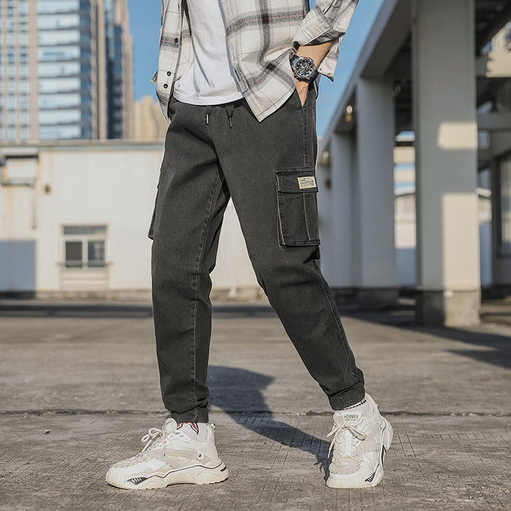 Un homme pose dans la rue avec une chemine à carreaux, un tee-shirt et des baskets blancs, une montre et un jean cargo noir resserré aux chevilles