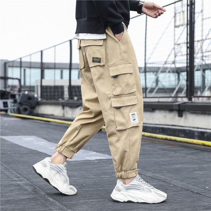 Un homme porte un pantalon cargo ample multipoches resserré aux chevilles beige de la marque Cargo District. Le pantalon est en coton 100% pour un confort maximal et une durabilité garantie. Il est disponible en différentes tailles, du S au 5XL.