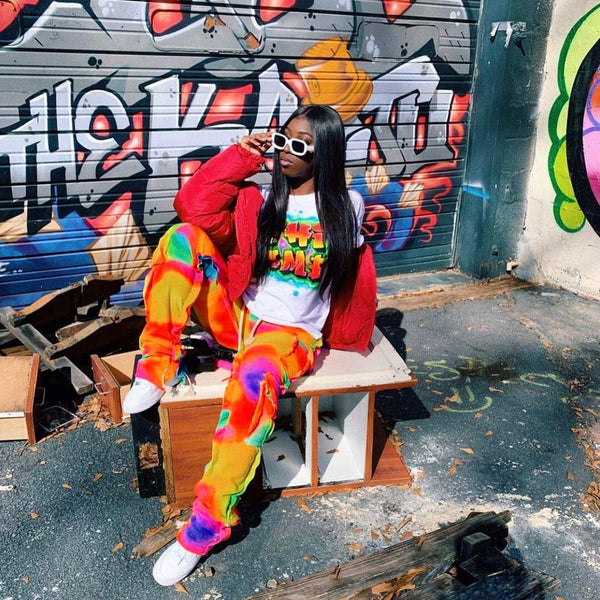 Une femme noire pose quand la rue devant des murs graffés. Elle porte un tee-shirt blanc à motif avec une doudoune rouge. Elle porte aussi des baskets blanches et un pantalon cargo jogger tie and dye multicolore. 