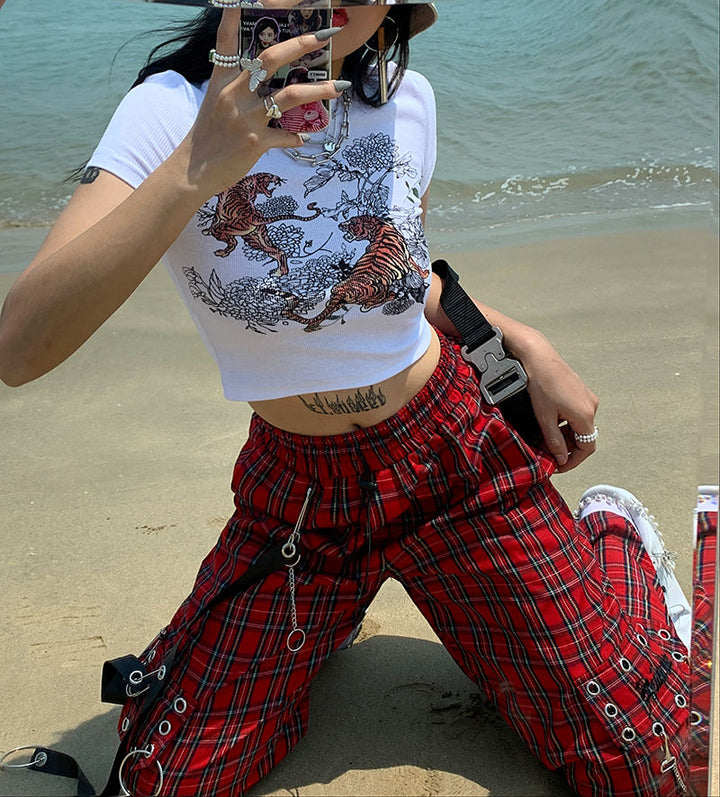 Une femme prenant un selfie sur une plage avec notre Pantalon cargo à carreaux et chaînes - Rouge - Femme. Le pantalon affiche un look rock avec sa coupe évasée et sa jambe large, orné d'une chaîne métallique, d'œillets en métal et de lanières en similicuir. Disponible du S au L.