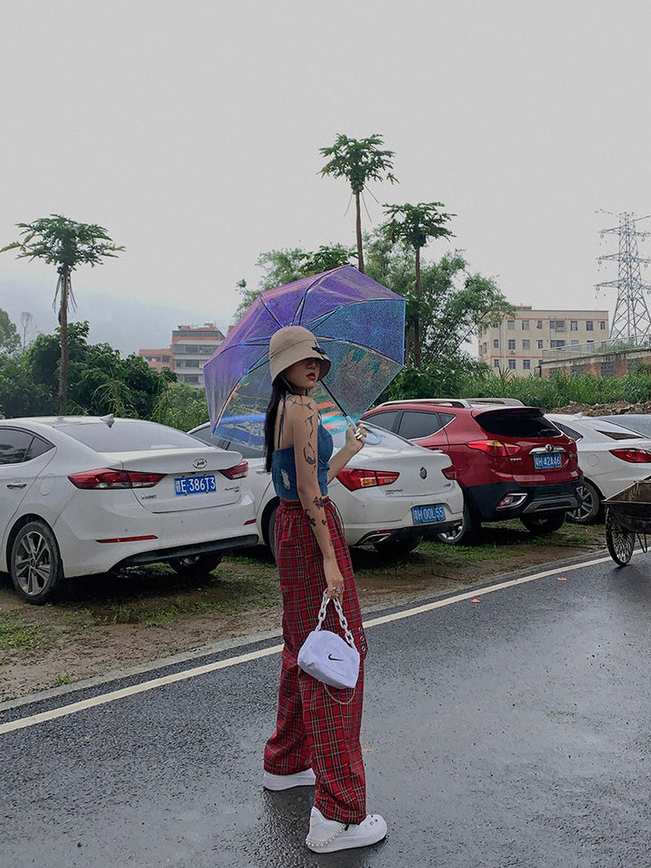 Une femme tenant un parapluie debout sur un parking, portant un pantalon cargo à carreaux et chaînes rouge. Le pantalon a une coupe évasée et une jambe large, avec des détails en métal et en similicuir. Disponible du S au L.