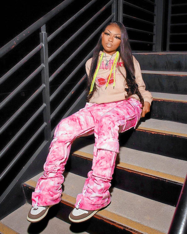 Une femme noire pose dans un escalier métallique. Elle porte un sweat beige à motif rose, des baskets blanches et marrons ainsi qu'un pantalon cargo jogger tie and dye rose.