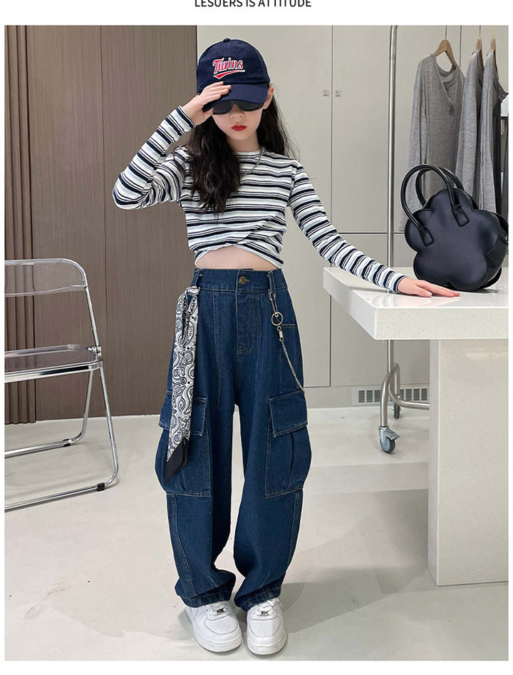 Une fille porte un pantalon cargo jean foulard chaîne bleu, exprimant sa personnalité avec style. Le pantalon a une coupe ample, une taille haute et des poches latérales pour plus de commodité. Disponible du 4 au 14 ans.