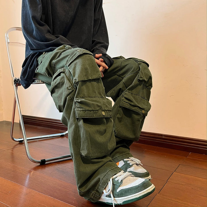 Un homme assis sur une chaise, portant un pantalon cargo baggy kaki avec de multiples poches. Look urbain et masculin pour se démarquer. Tailles disponibles de M à 2XL. Confortable et durable en 100% coton.