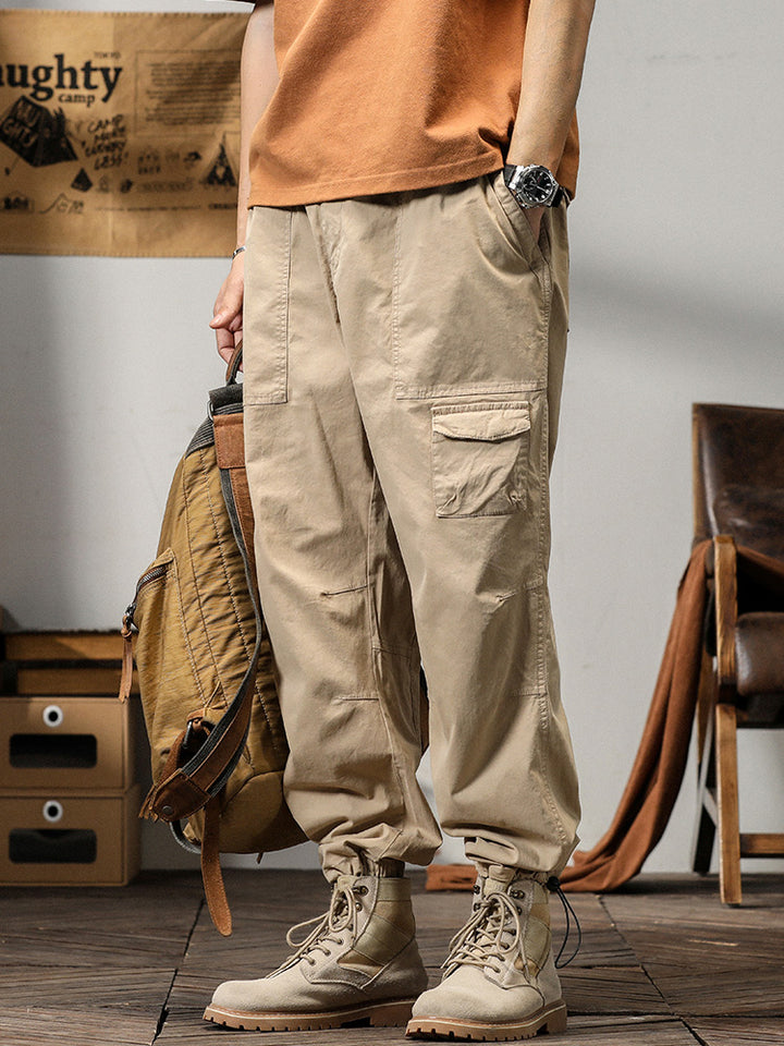 Un homme porte un pantalon cargo léger ample coupe droite beige avec un sac à dos. Le pantalon est confortable et flexible, idéal pour l'été. Les poches sur les côtés ajoutent une touche fonctionnelle. Disponible du M au 3XL.