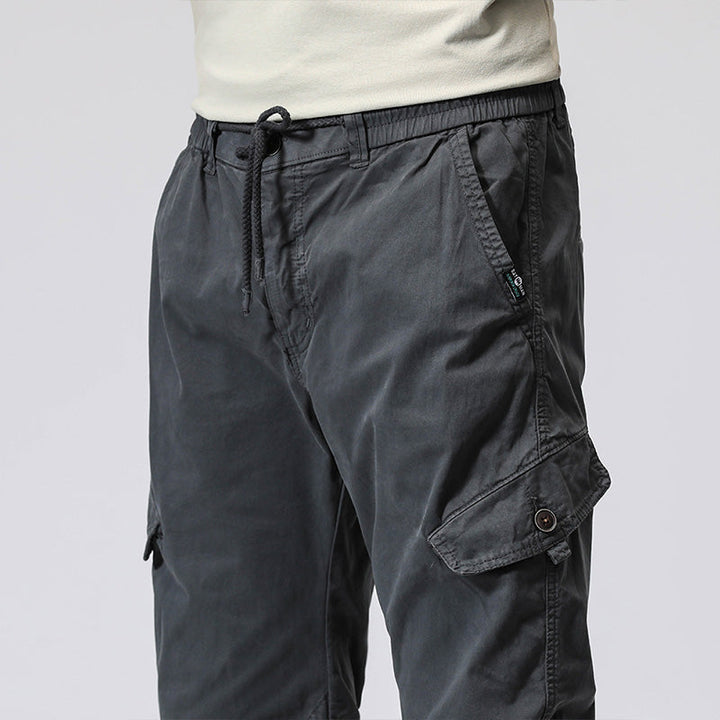 Un homme portant un pantalon cargo gris avec fermeture rouge, coupe droite et resserrée à la cheville. Le pantalon est composé de 95% coton et 5% spandex, offrant douceur, résistance et souplesse. Disponible du 28 au 38 (tailles US).