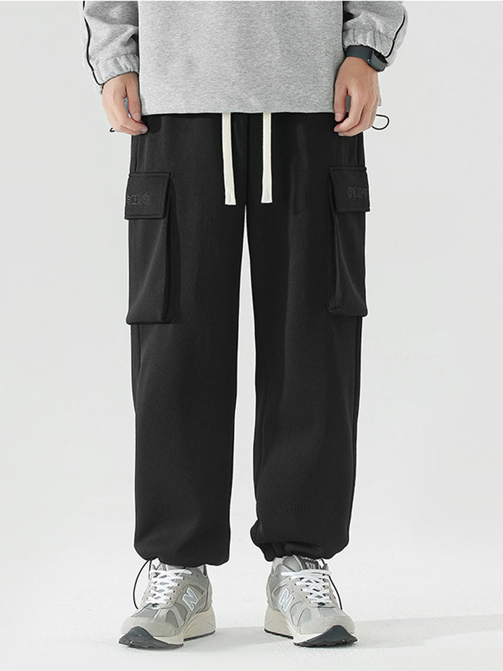 Un homme porte un pantalon cargo baggy noir avec une inscription cousue sur une poche latérale. Le pantalon offre une liberté de mouvement optimale grâce à son ampleur et à l'élastique à la taille et à la cheville. Fabriqué en 100% polyester, il est résistant et durable. Disponible du S au 2XL.