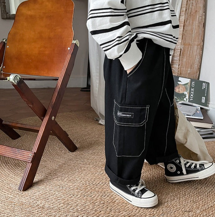 Un garçon se tient à côté d'une chaise en bois pliante avec un cadre en métal. Il porte un pantalon cargo noir avec des coutures contrastantes blanches. Un magazine et un ordinateur portable sont posés sur une table à côté de lui.