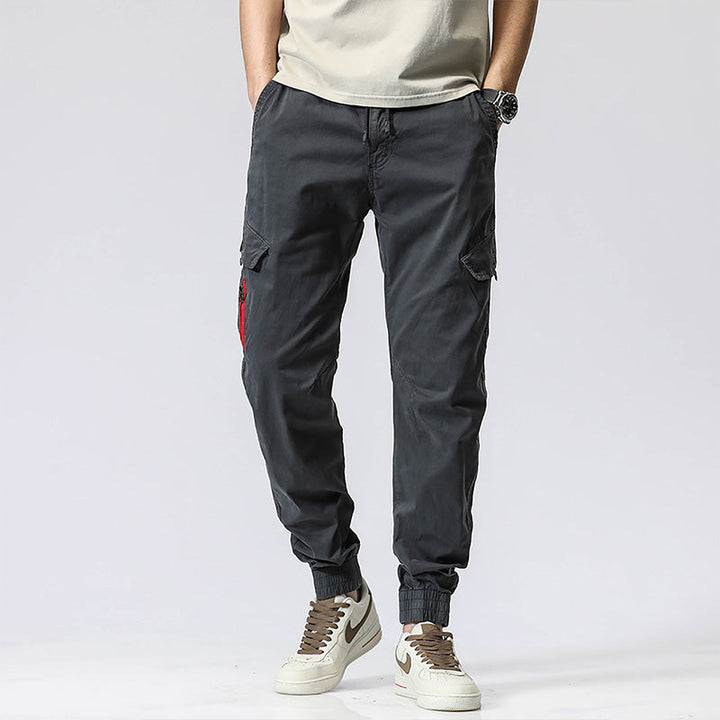 Un homme porte un pantalon cargo gris avec une fermeture rouge sur une poche. Coupe droite et resserrée à la cheville, ce pantalon cargo est à la fois élégant et décontracté. Disponible en différentes tailles du 28 au 38 (tailles US). Matières : 95% coton et 5% spandex. Idéal pour un style cargo.