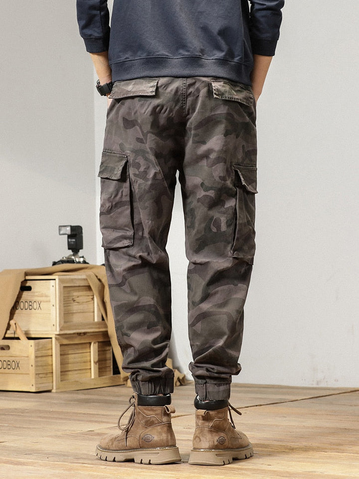 Un homme portant un pantalon cargo gris camo avec coupe large resserrée aux chevilles. Style militaire masculin et moderne.