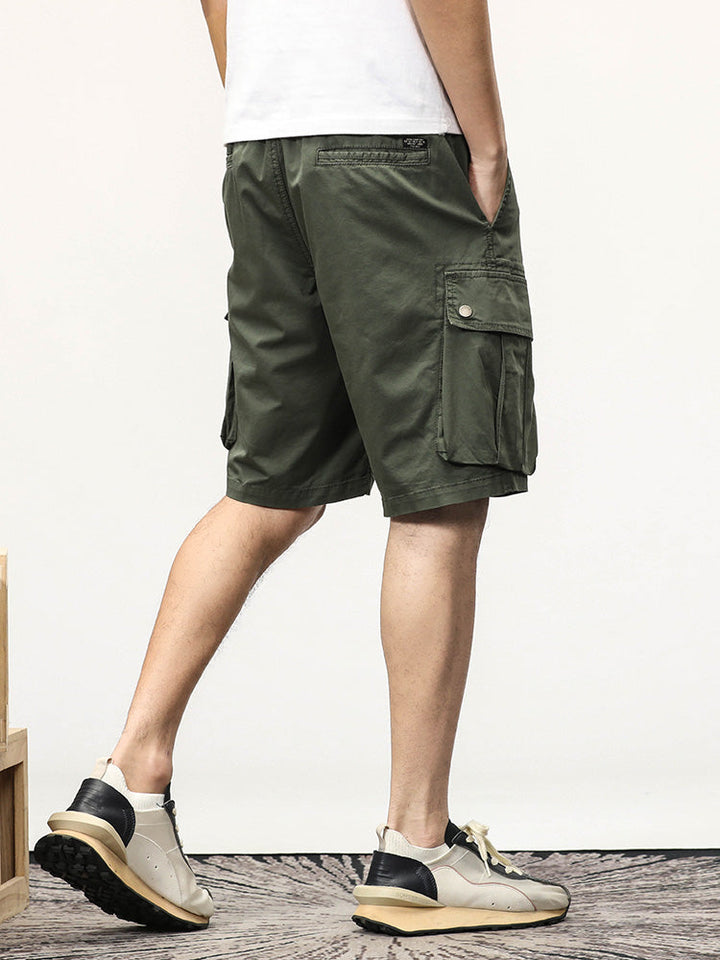 Un homme porte un short cargo coupe droite kaki avec des sneakers. Le short a des poches latérales à boutons-pression et une fermeture élastique avec un cordon à la taille. Le short est fabriqué à partir de 95% coton et 5% spandex pour un confort optimal. Disponible en tailles US du 28 au 40.