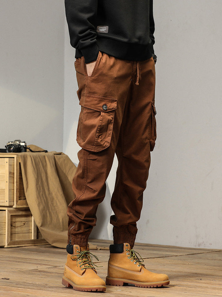 Un homme portant un pantalon cargo coupe droite avec poches à bouton - Camel. Le pantalon a des poches à rabats avec bouton apparent pour une fermeture sécurisée. Coupe droite avec bas resserré aux chevilles. Tailles disponibles du 28 au 38.