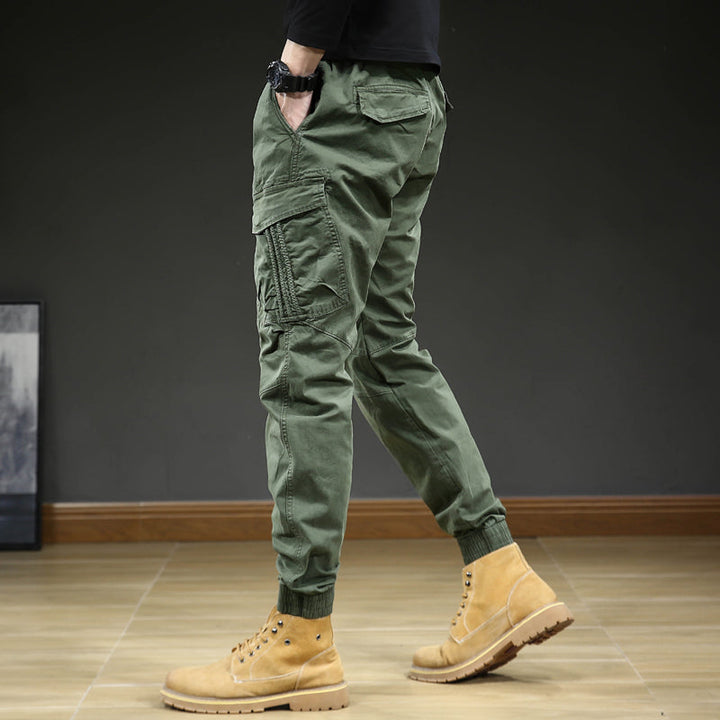 Un homme porte un pantalon cargo slim resserré aux chevilles de couleur kaki. Le pantalon est confortable et élégant avec une coupe slim et des coutures aux genoux. Disponible en tailles US 28 à 38.