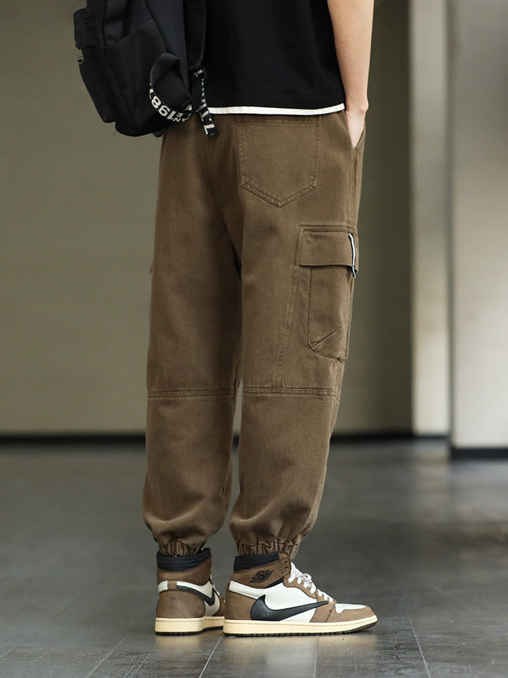 Un homme porte un pantalon cargo ample resserré aux chevilles de couleur marron clair. Le pantalon est doté de poches cargo latérales et est fabriqué à partir d'un mélange de coton et de viscose pour plus de douceur et de confort. Disponible dans différentes tailles.