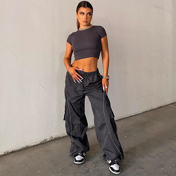 Une femme pose dans un parking avec un tee-shirt moulant et court gris foncé, des baskets noires et blanches. Elle porte aussi un pantalon jogging cargo baggy gris foncé. Il est taille haute. La femme porte une queue de cheval et des bijoux. 