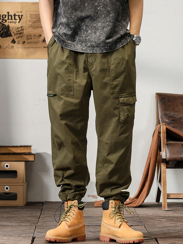 Un homme pose dans un décor industriel avec un tee shirt gris, une montre et des boots marrons. Il porte également un pantalon cargo coupe droite ample et léger kaki.