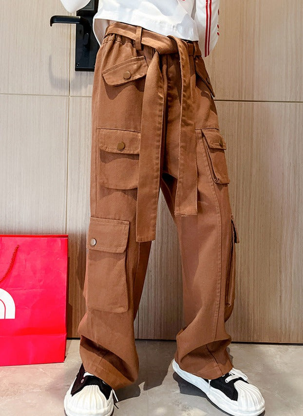 Une personne portant un pantalon cargo marron taille haute avec ceinture - Marron - Fille, avec des poches latérales. Confortable et à la mode, ce pantalon cargo large est un incontournable pour votre petite fille. Disponible du 120 au 170 cm.