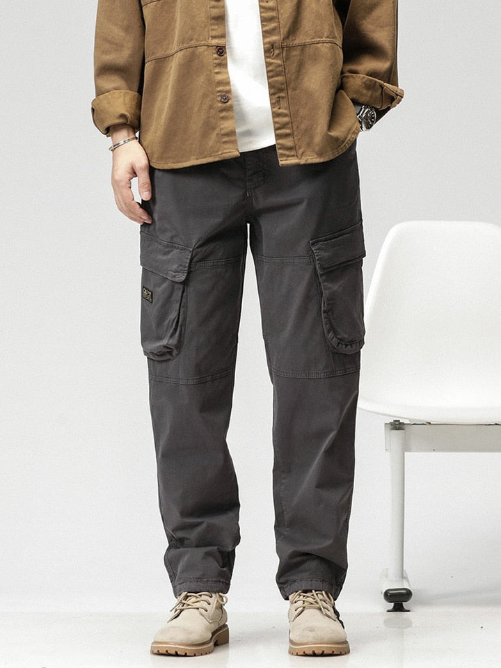 Un homme pose debout devant une chaise blanche. Il porte un tee-shirt blanc avec une chemise ouverte camel, des boots beige. Il porte aussi un pantalon cargo ample coupe droite gris.