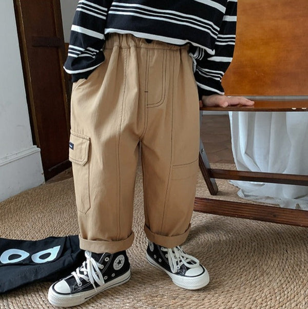 Un enfant pose dans un salon. Il y a un tapis rond en jute, une chaise. L'enfant porte une marinière noire à rayures blanches, des baskets montantes noires et blanches. Il porte un pantalon cargo beige ample avec des coutures contrastantes noires.