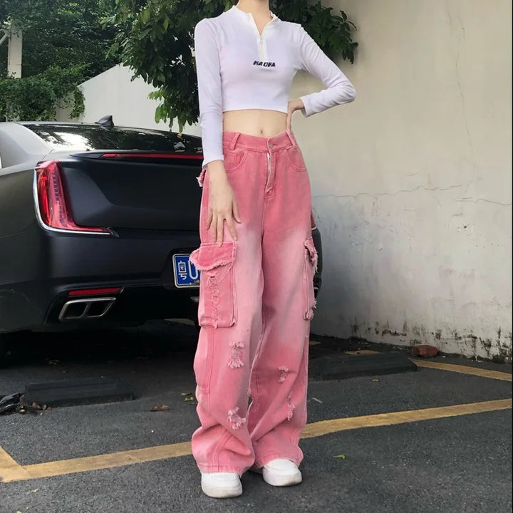 Une femme debout devant une voiture, portant un jean cargo baggy rose délavé déchiré taille haute. Un look audacieux des années 2000 avec une touche d'insouciance et de féminité. Disponible en tailles XS à XL. Revisitez le style cargo avec Cargo District.