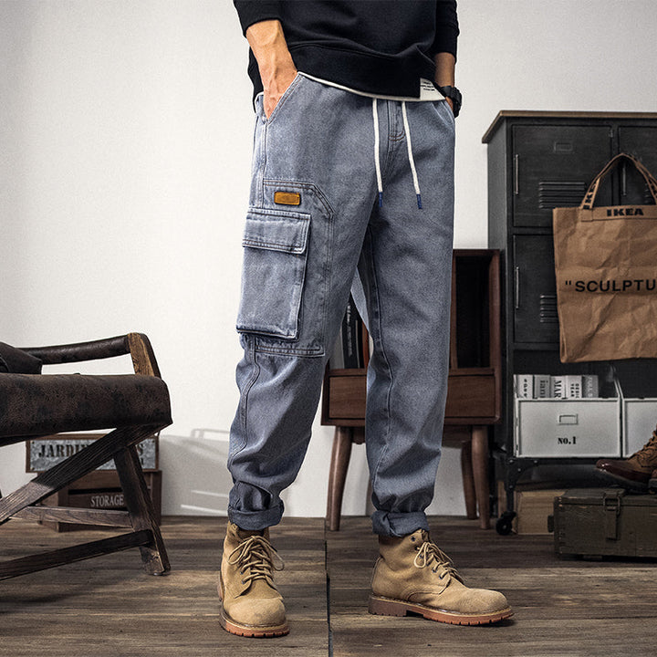 Un homme porte un jean cargo coupe large bleu clair avec des poches latérales cargo. Le jean est confectionné en coton et polyester pour un vêtement robuste et confortable. Tailles disponibles du XS au 5XL.