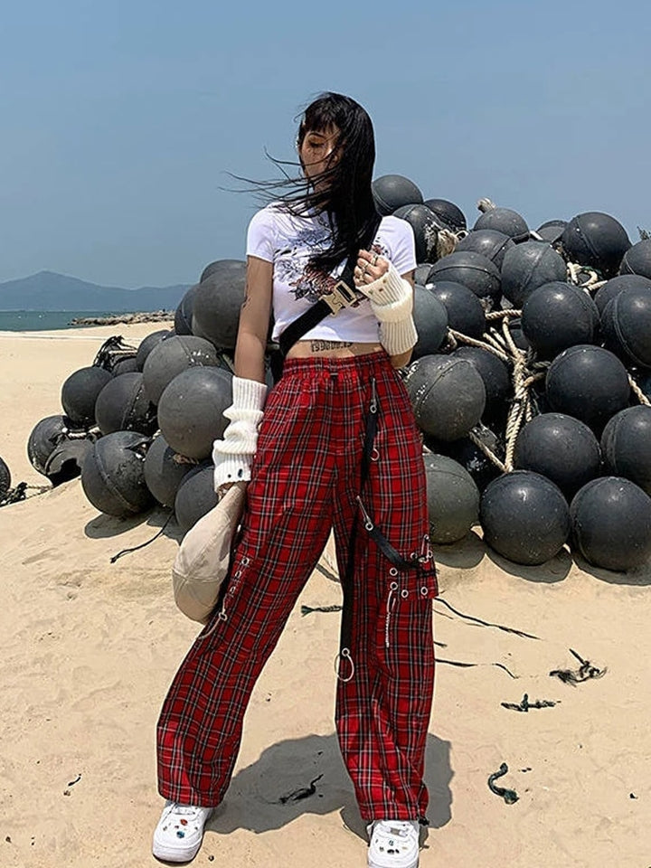 Une femme debout devant un tas de balles noires, portant un Pantalon cargo à carreaux et chaînes rouge. Le pantalon taille haute affiche un look rock avec sa coupe évasée et sa jambe large. Orné d'une chaîne métallique, d’œillets en métal et de lanières en similicuir, ce pantalon arbore également des épingles sur les côtés pour une touche décalée. Disponible du S au L.