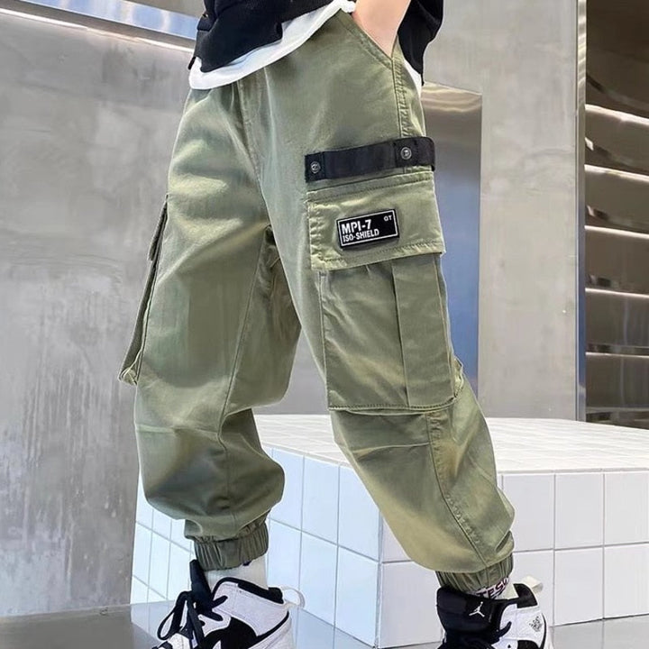 Un enfant portant un pantalon cargo jogger large kaki avec des poches latérales pratiques. Le pantalon est resserré aux chevilles et a une taille élastique pour un ajustement parfait et un confort maximal. Disponible du 110 au 160 cm.