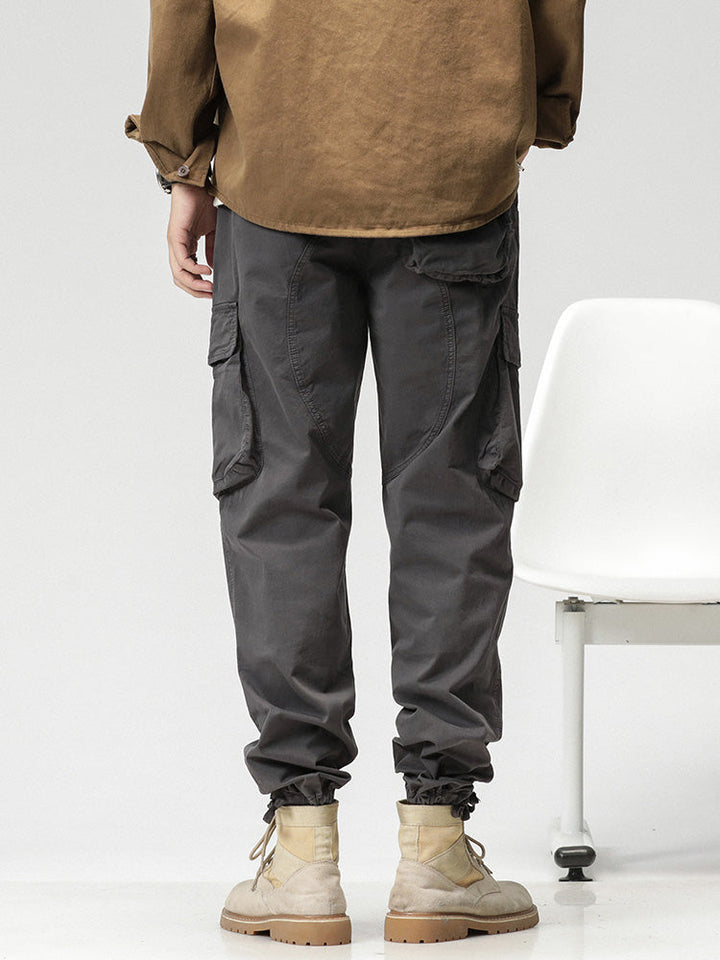 Un homme porte un pantalon cargo ample coupe droite gris avec des poches latérales et une fermeture par bouton et fermeture éclair. Le pantalon en coton et spandex offre confort et flexibilité. Les jambes peuvent être resserrées aux chevilles pour un look sportif ou portées droites pour un look classique. Disponible du 28 au 38 (tailles US).