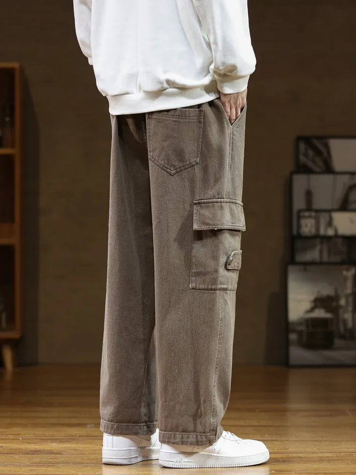 Un homme porte un pantalon cargo ample en coton marron. Ce pantalon est résistant et offre une grande liberté de mouvement grâce à sa coupe ample. La ceinture intégrée ajoute une touche utilitaire chic. Disponible dans une large gamme de tailles, du M (FR36) au 8XL (FR56).