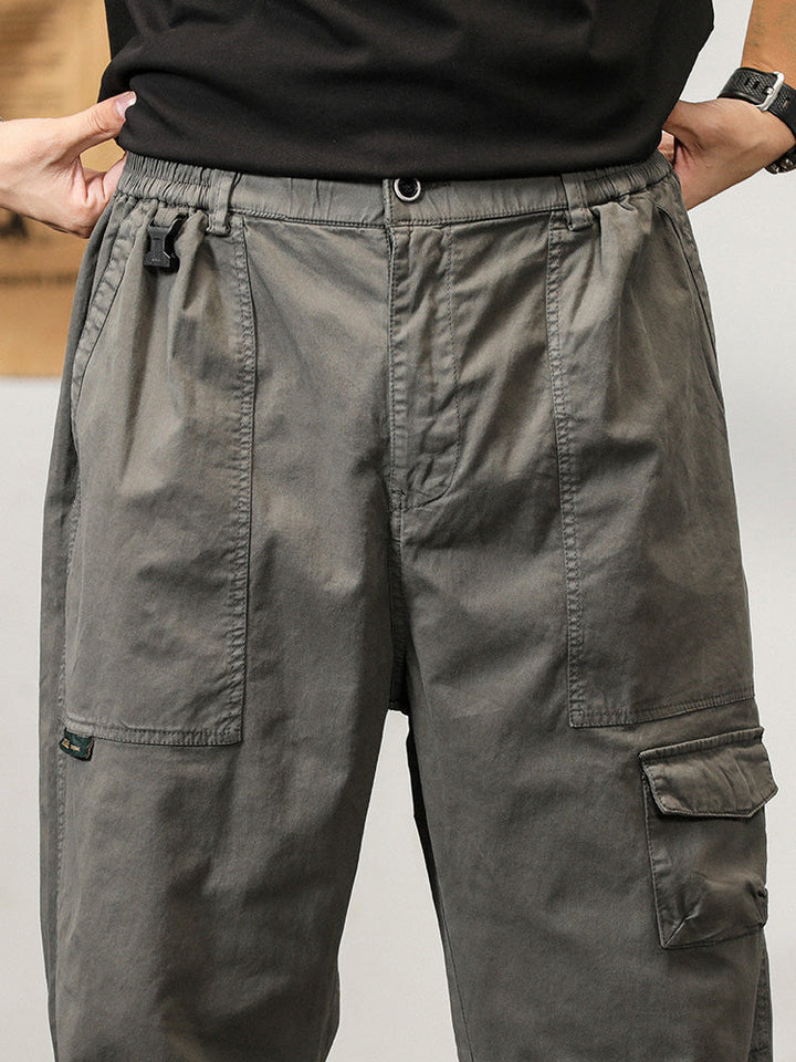 Un homme porte un pantalon cargo léger ample coupe droite gris avec des poches fonctionnelles. Parfait pour l'été, ce pantalon offre confort et maintien grâce à l'élastique à la taille et la fermeture par bouton et fermeture éclair. Disponible du M au 3XL.