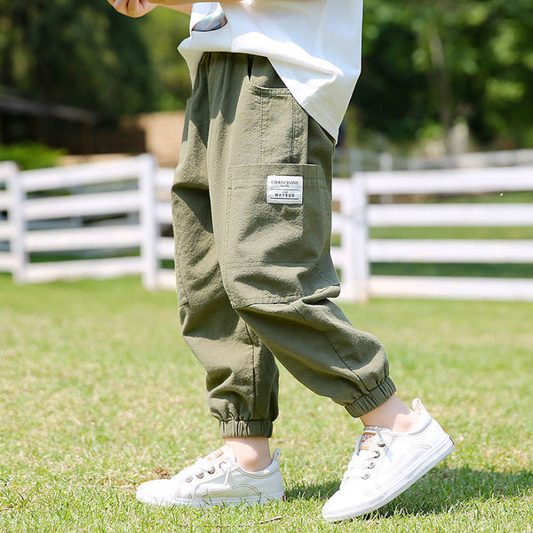 Un enfant pose dans un jardin avec un clotûre blanche dans le fond. Il porte un tee-shirt blanc, des baskets blanches ainsi qu'un pantalon cargo jogger en coton kaki. 