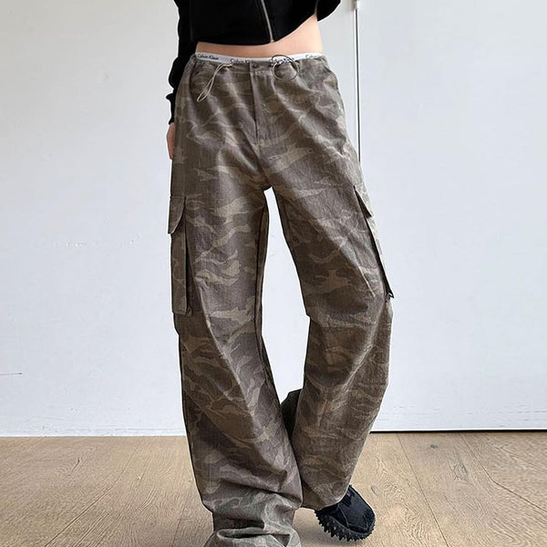 Une femme pose dans une pièce aux murs blanc et parquet au sol. Elle porte un gilet noir, des baskets noirs ainsi qu'un pantalon cargo baggy taille basse avec élastique à la taille vert camo.