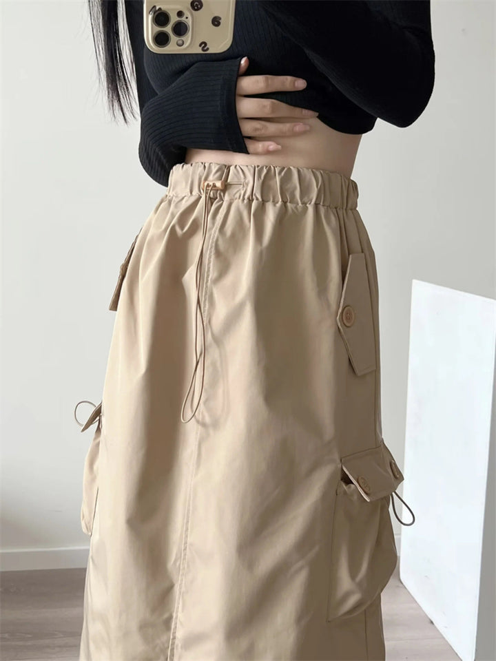 Une femme portant une jupe cargo longue légère beige avec taille haute et fente arrière. Poches boutonnées pour un look utilitaire tendance. Disponible du S au L.