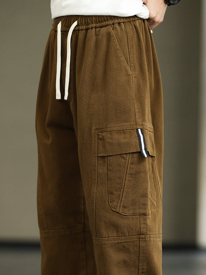 Un homme porte un pantalon cargo ample resserré aux chevilles de couleur marron. Le pantalon est doté de poches cargo latérales et d'une ceinture élastique avec cordon pour un ajustement parfait. Fabriqué en 95% coton et 5% viscose, il offre douceur, confort et durabilité. Disponible du M au 8XL. Parfait pour un style street tendance.