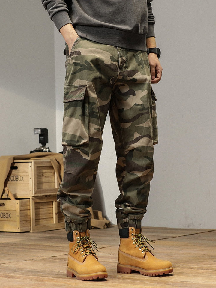 Un homme pose dans une pièce avec parquet au sol et murs blancs. Il y a des boîtes en bois et un appareil photo derrière lui. Il porte un sweat gris, des boots camel et un pantalon cargo ample resserré aux cheviles kaki camo.