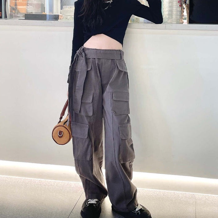 Une femme appuyée contre un mur, portant un Pantalon cargo large gris taille haute avec ceinture - Fille. Multiples poches latérales, coupe large à la mode. Confortable et stylé. Disponible du 120 au 170 cm.