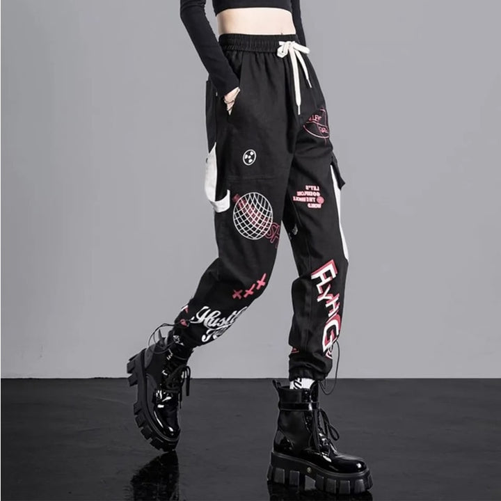 Une personne portant un pantalon cargo jogger noir à motifs avec des bottes noires. Le pantalon cargo noir présente une coupe jogger taille haute avec des motifs blancs et roses pour une allure urbaine. Il est doté d'une poche latérale et d'une lanière gris clair pour une touche d'originalité. Disponible du S au 3XL, ce pantalon en coton offre un confort optimal.