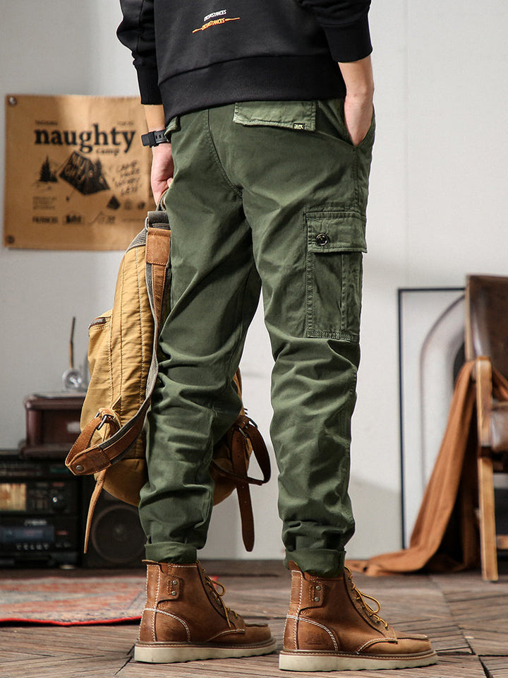 Un homme en pantalon cargo kaki tenant un sac à dos. Coupe droite, poches cargo sur les côtés. Confortable et stylé. Disponible en tailles US 28 à 38.