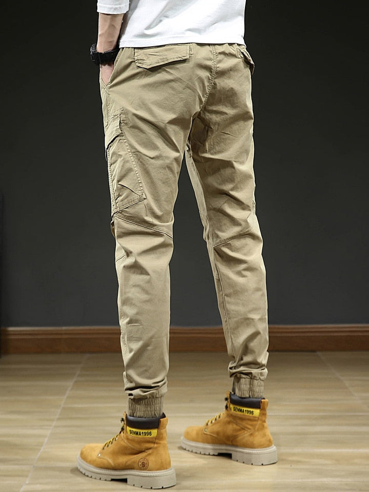 Un homme porte un pantalon cargo slim resserré aux chevilles de couleur beige avec des bottes jaunes. Le pantalon est confortable et élégant, avec une coupe slim et des coutures aux genoux. Disponible en tailles US 28 à 38.