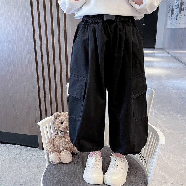 Une enfant pose sur une chaise avec un pull blanc, des baskets blanches, des chaussettes roses et un pantalon cargo baggy noir. Il y a un nounours à côté d'elle.