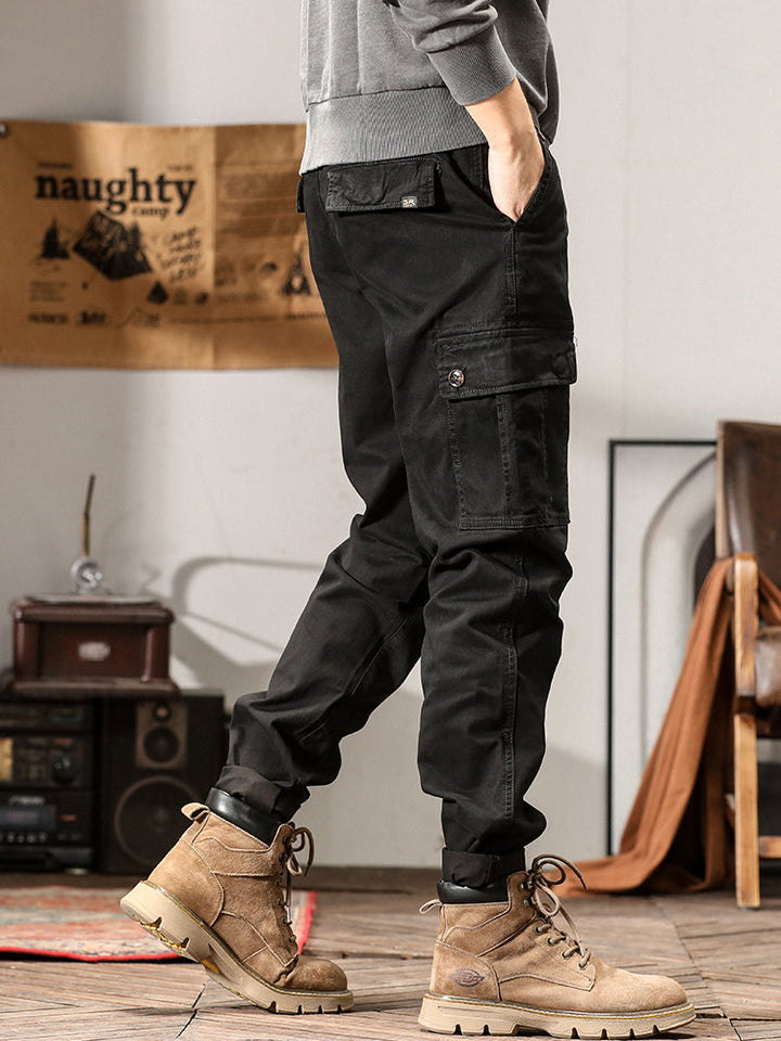 Un homme porte un pantalon cargo noir avec des bottes. Le pantalon a une coupe droite et des poches cargo sur les côtés. Idéal pour une variété d'occasions.