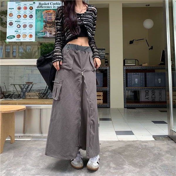 Une femme pose devant un restaurant avec une jupe longue cargo grise évasée. La jupe est ornée d'un zip vertical sur le devant de la jupe. La femme porte également un sac noir, un crop top noir ainsi qu'un gilet court ouvert noir et blanc. Elle porte aussi des baskets noires et blanches.