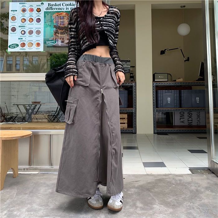 Une femme pose devant un restaurant avec une jupe longue cargo grise évasée. La jupe est ornée d'un zip vertical sur le devant de la jupe. La femme porte également un sac noir, un crop top noir ainsi qu'un gilet court ouvert noir et blanc. Elle porte aussi des baskets noires et blanches.