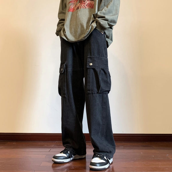 Un homme pose dans une pièce vide aux murs beige et parquet au sol. Il porte un pull gris à motif, des basket noires et blanches ainsi qu'un jean cargo baggy noir. 